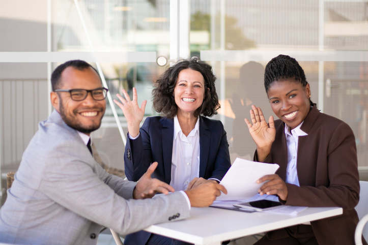 Happy successful business team posing at table and waving hello. Cheerful multiethnic man and women sitting in outdoor cafe and smiling at camera. Team success concept.
