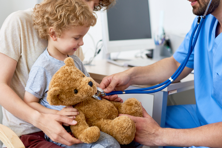 Portrait of adorable curly child sitting on mothers lap in doctors office holding teddy bear toy, with pediatrician listening to heartbeat using stethoscope. child getting checked for hydroceles